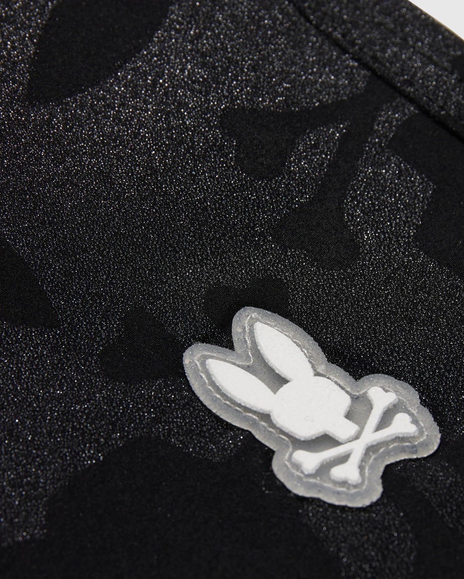 Close-up of a textured black fabric with a subtle pattern, featuring a white Playboy bunny logo appliqué on Psycho Bunny MENS MALTA HYDROCHROMIC SWIM TRUNK - B6W321B2SW.
