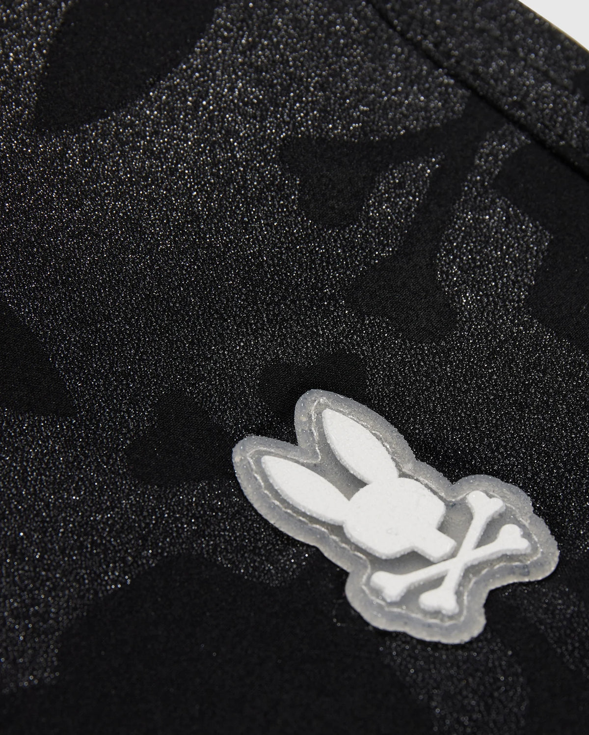 Close-up of a textured black fabric with a subtle pattern, featuring a white Playboy bunny logo appliqué on Psycho Bunny MENS MALTA HYDROCHROMIC SWIM TRUNK - B6W321B2SW.