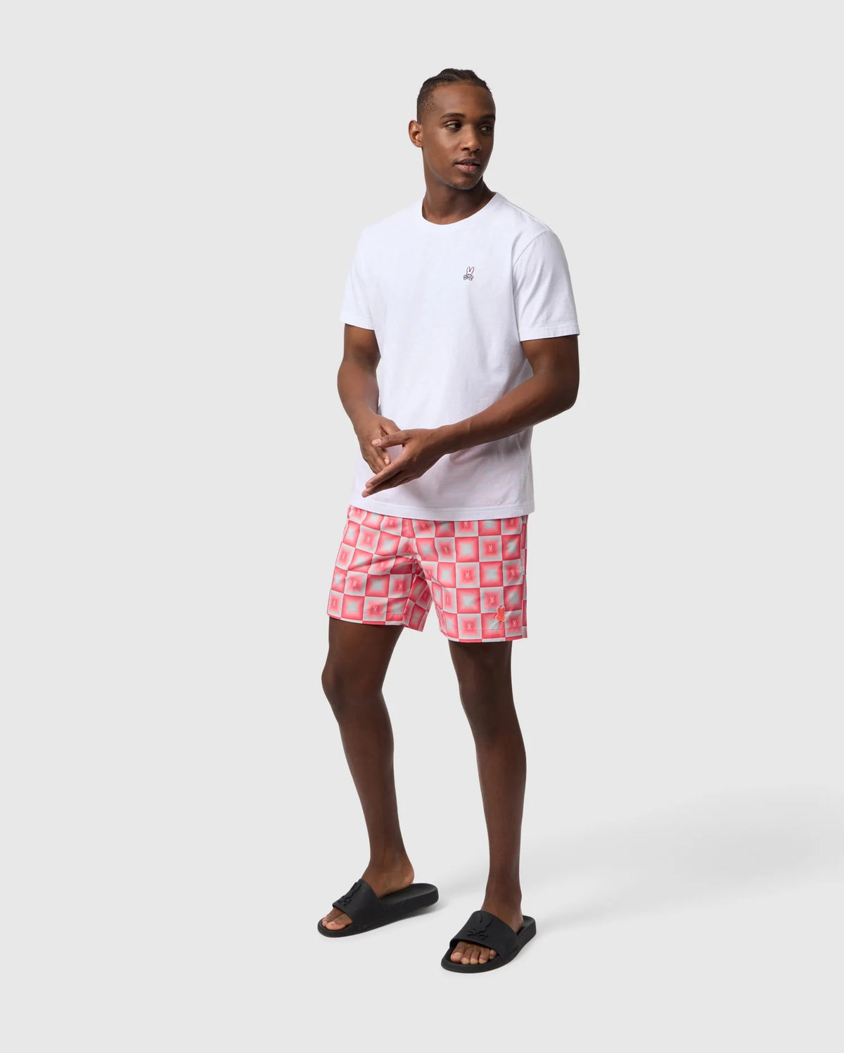 A man in a white t-shirt and red Psycho Bunny UTICA AOP swim trunk stands in a neutral stance against a light gray background, looking to the side. He wears black flip-flops.