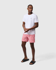 A man in a white t-shirt and red Psycho Bunny UTICA AOP swim trunk stands in a neutral stance against a light gray background, looking to the side. He wears black flip-flops.