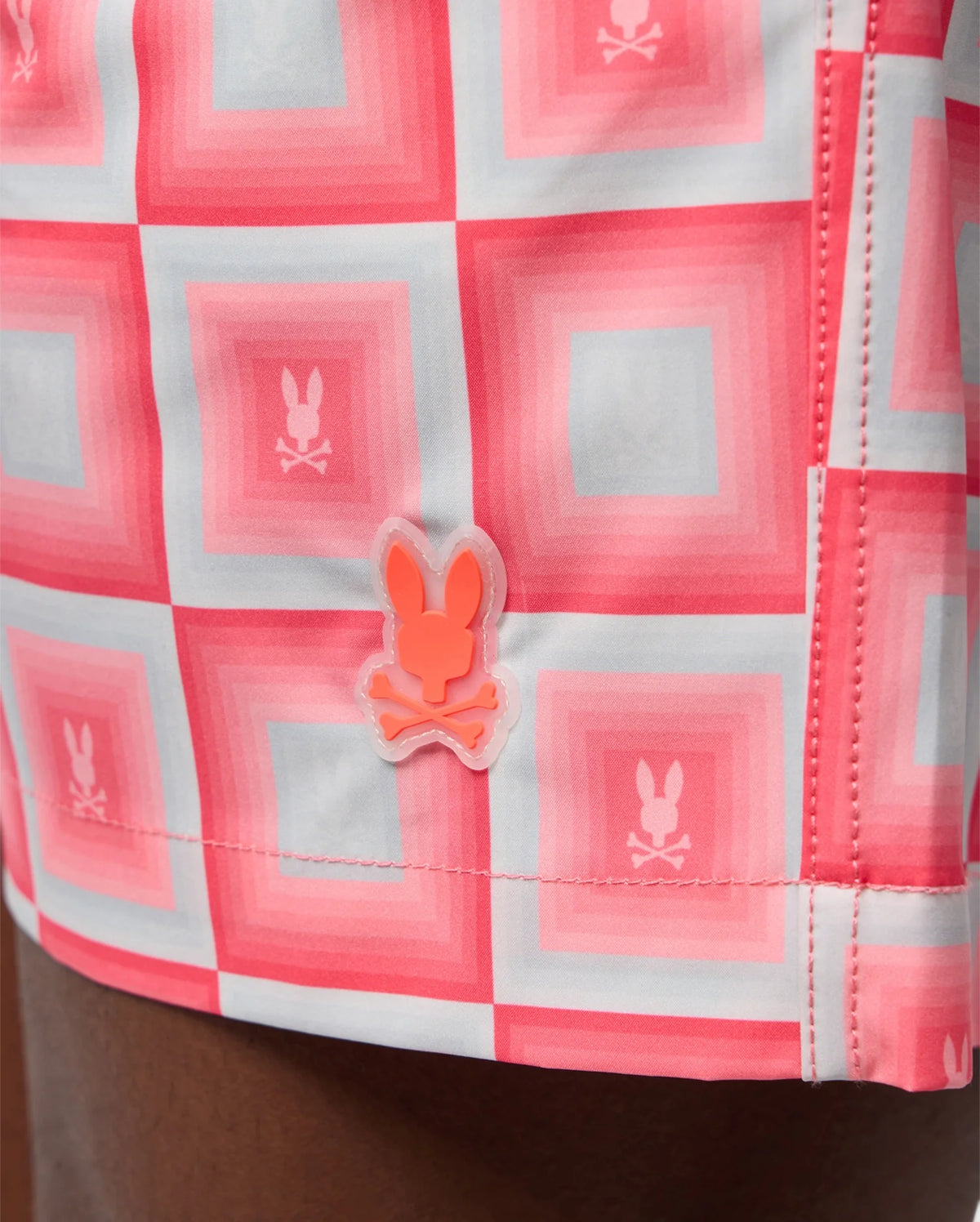 Close-up of a Psycho Bunny MENS UTICA SWIM TRUNK - B6W399B2SW with a red and pink grid pattern featuring repeated bunny logos, and a bunny-shaped pin attached to it.
