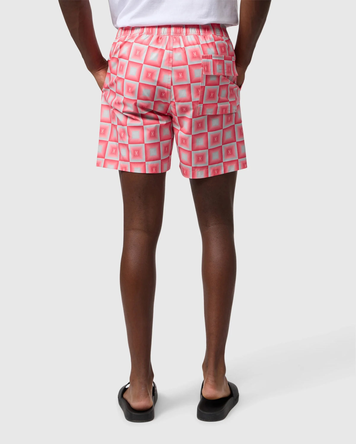 A person from the waist down wearing Psycho Bunny MENS UTICA SWIM TRUNK - B6W399B2SW with a repeated pattern of white squares and red x’s, paired with black flip-flops, standing against a white background.