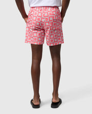 A person from the waist down wearing Psycho Bunny MENS UTICA SWIM TRUNK - B6W399B2SW with a repeated pattern of white squares and red x’s, paired with black flip-flops, standing against a white background.