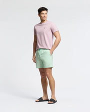 A person stands against a plain background wearing a light pink t-shirt, Psycho Bunny mint green swim trunks (MENS SAMOS HYDROCHROMIC PRINTED SWIM TRUNK - B6W453F200) with subtle bunny pattern, and black sandals. They have one hand in their pocket and look to the side.