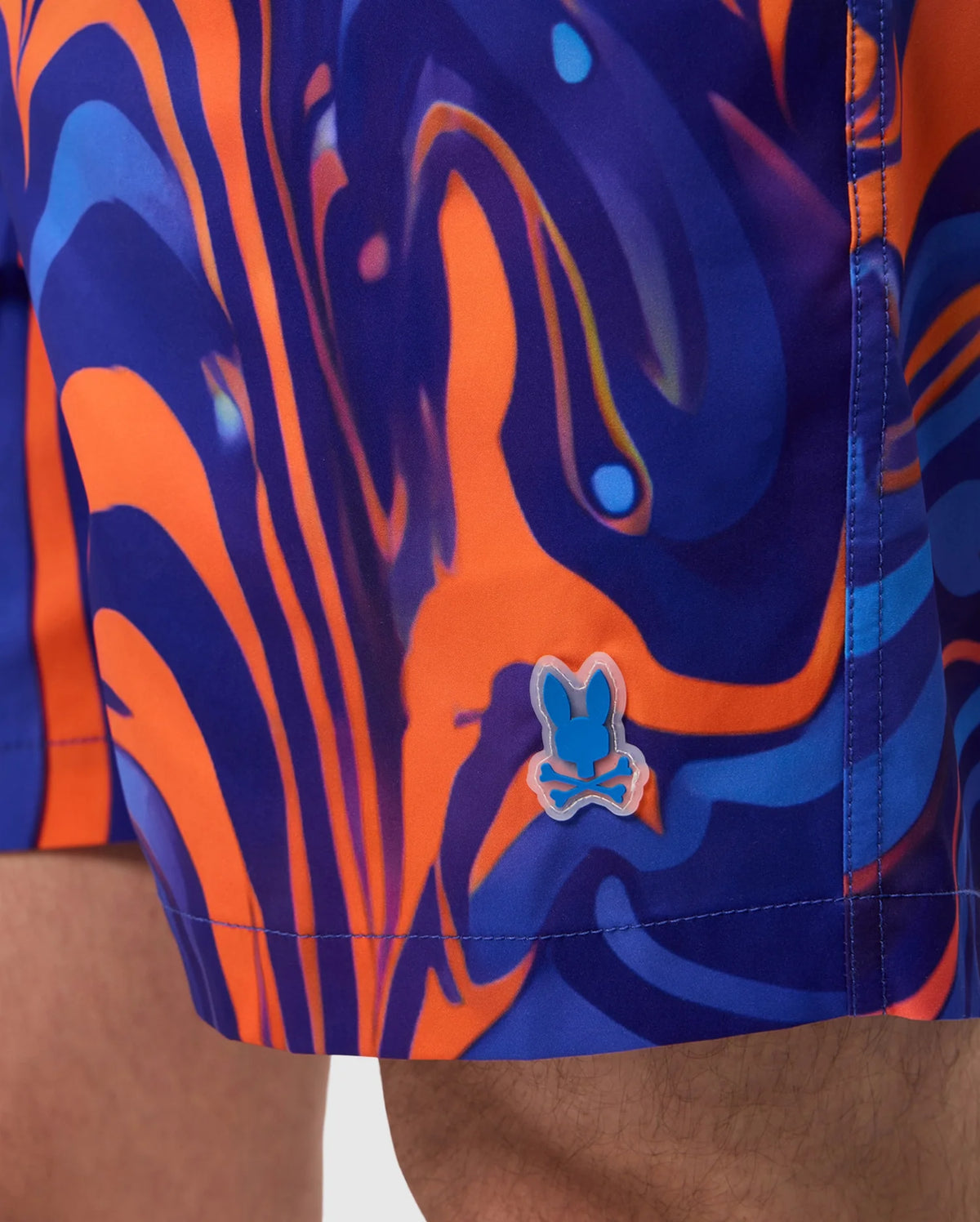 Close-up of a person wearing Psycho Bunny MENS NEVADA ALL OVER PRINT SWIM TRUNK - B6W526C200 featuring an abstract swirl print in orange and blue hues. Made from quick-dry fabric, the design showcases swirling lines and shapes. A small emblem of a stylized rabbit skull and crossbones is visible near the hem.