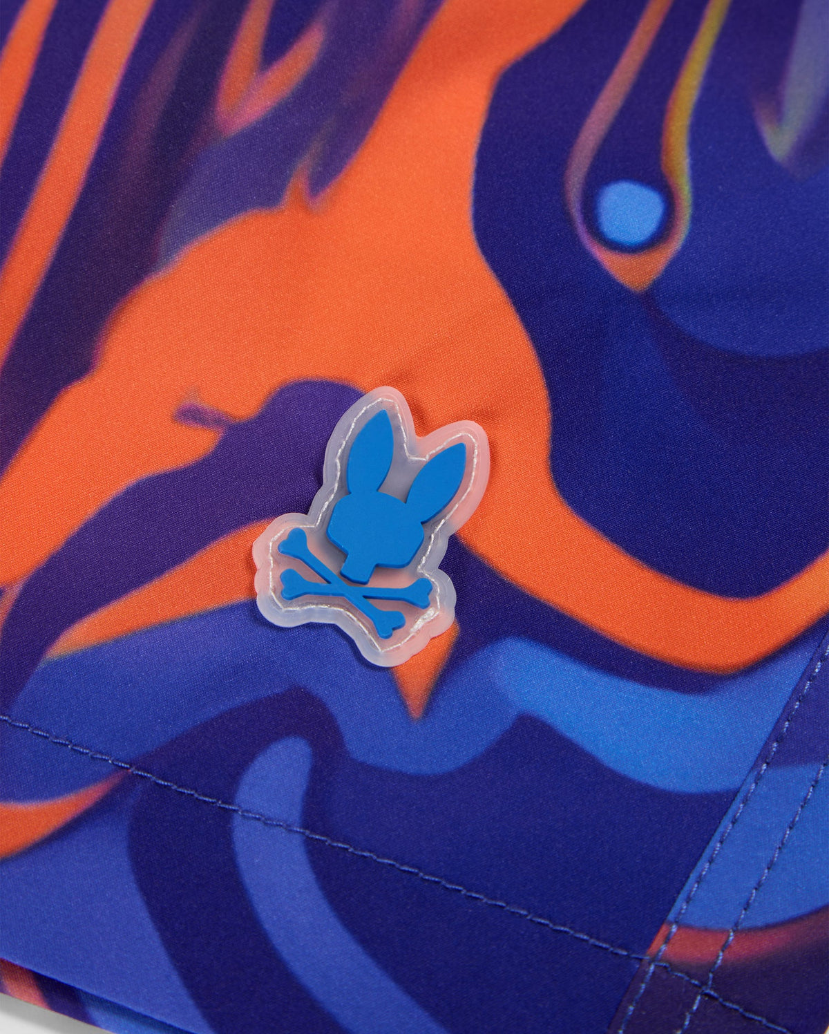 Close-up of a colorful fabric featuring a blend of orange, blue, and purple abstract patterns. The image also shows a small rubber emblem of a bunny skull and crossbones in blue and white, affixed to the vibrant swirl print of Psycho Bunny MENS NEVADA ALL OVER PRINT SWIM TRUNK - B6W526C200.