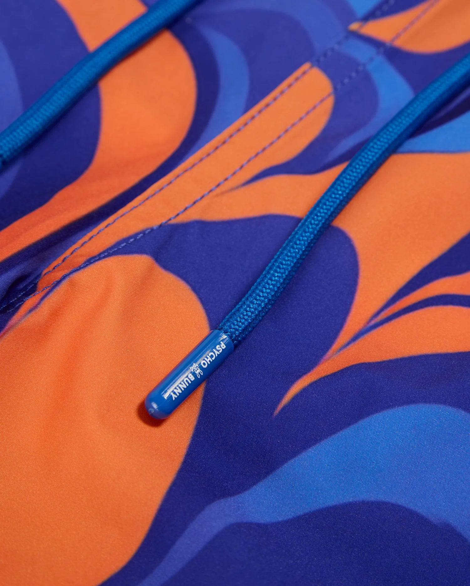Close-up of a vibrant swirl print on bright orange and blue patterned swim trunks, featuring abstract swirled designs. A blue drawstring with a plastic tip, labeled with small white text, is partially visible in the frame. The quick-dry fabric's stitching and drawstring tunnel are also clearly defined. These are the MENS NEVADA ALL OVER PRINT SWIM TRUNK - B6W526C200 by Psycho Bunny.