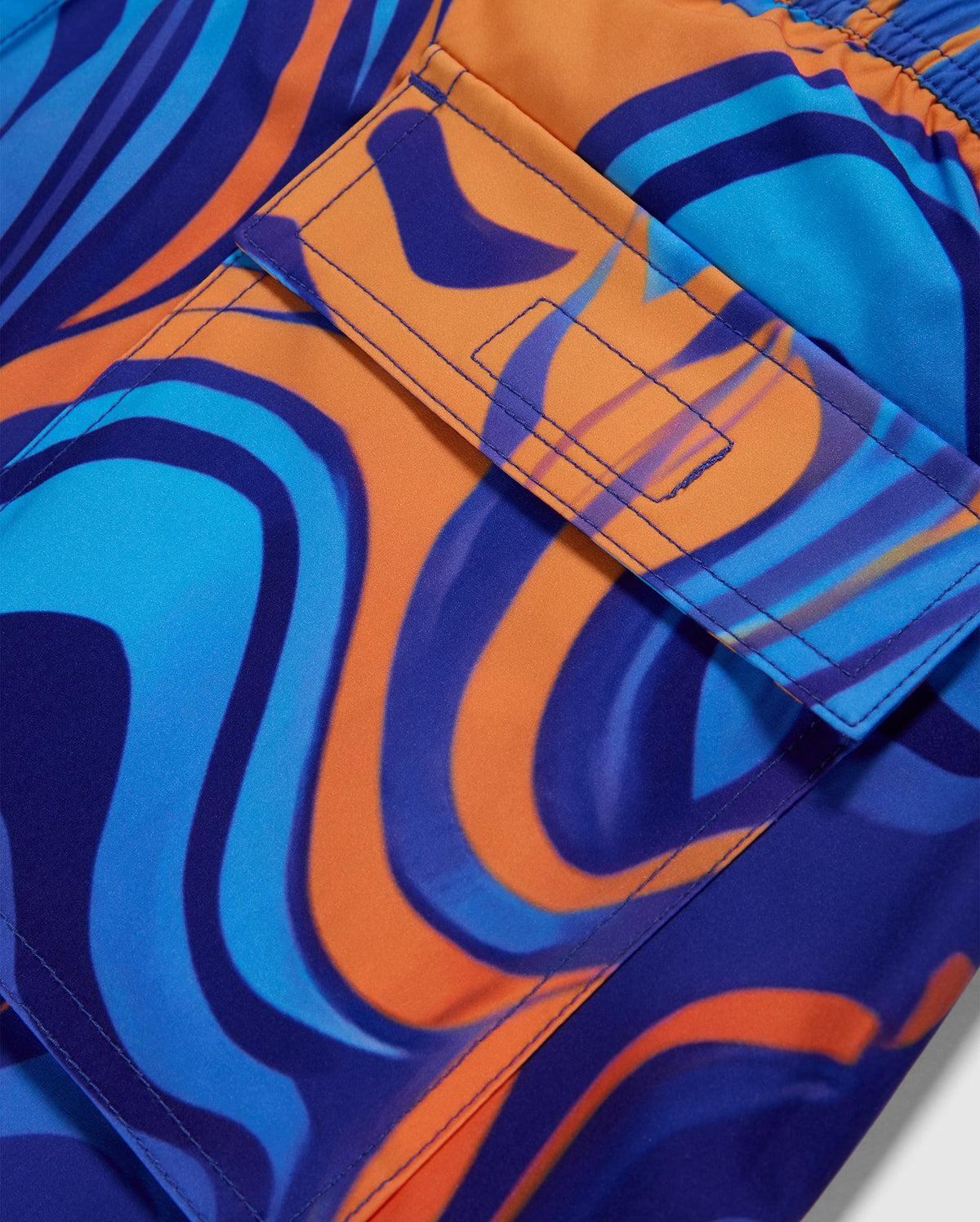Close-up of Psycho Bunny MENS NEVADA ALL OVER PRINT SWIM TRUNK - B6W526C200 featuring a vibrant swirl print in blue, orange, and purple shades. The quick-dry fabric includes a pocket with a flap secured by a Velcro strip, showcasing intricate stitching and clean lines. The pattern resembles fluid, wavy shapes and gradients.