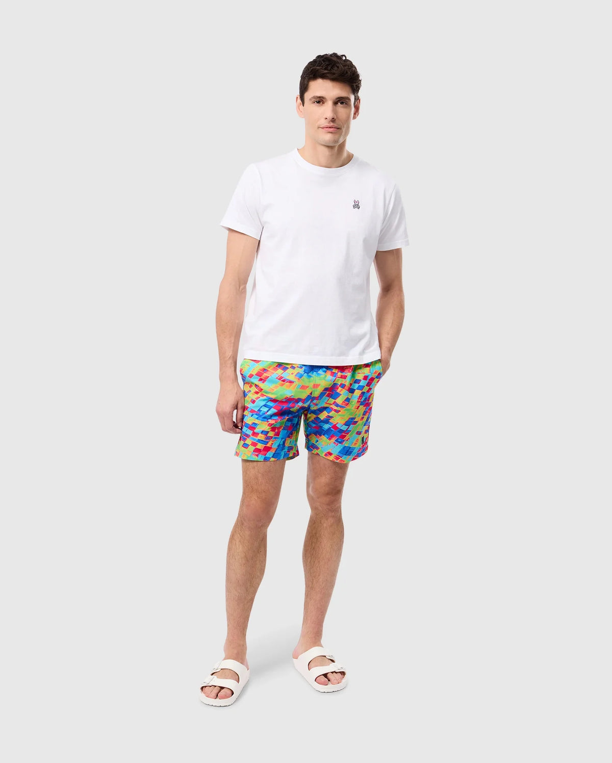 A man is standing against a plain white background, wearing a white T-shirt, MENS DENNIS ALL OVER PRINT SWIM TRUNK - B6W530C200 from Psycho Bunny made from quick-dry fabric, and white slide sandals. His hands are relaxed, with one hand in his pocket.