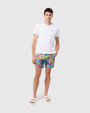 A man is standing against a plain white background, wearing a white T-shirt, MENS DENNIS ALL OVER PRINT SWIM TRUNK - B6W530C200 from Psycho Bunny made from quick-dry fabric, and white slide sandals. His hands are relaxed, with one hand in his pocket.
