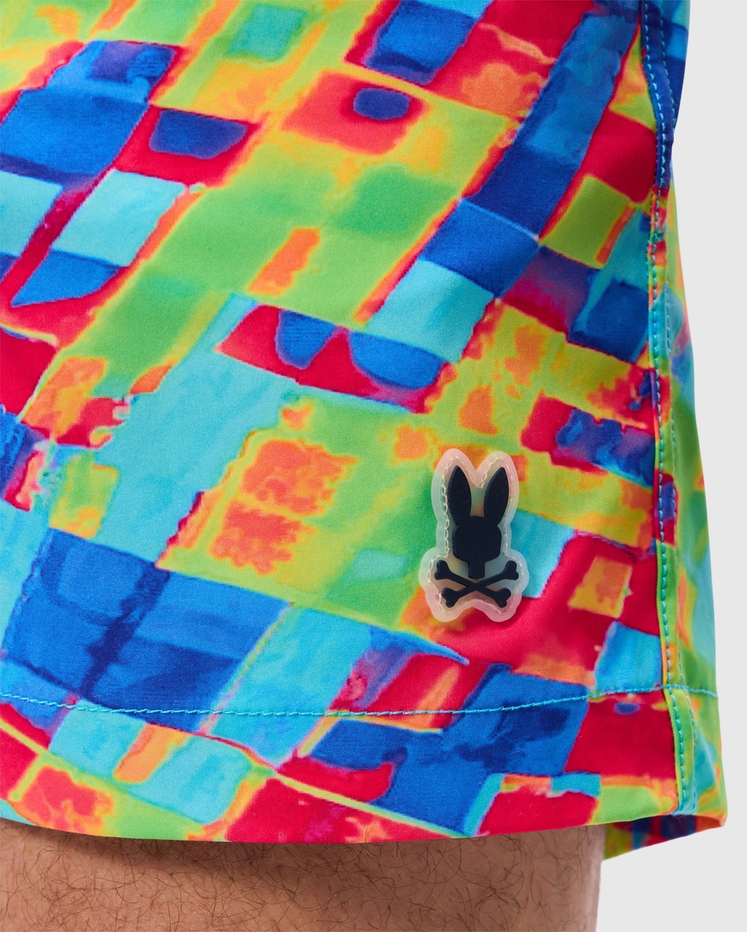 A close-up of the MENS DENNIS ALL OVER PRINT SWIM TRUNK - B6W530C200 by Psycho Bunny, showcasing its vibrant geometric pattern in red, green, blue, and yellow. Made from quick-dry fabric, the trunks feature a small black and white bunny logo near the hem. A portion of a person's leg is visible at the bottom of the image.