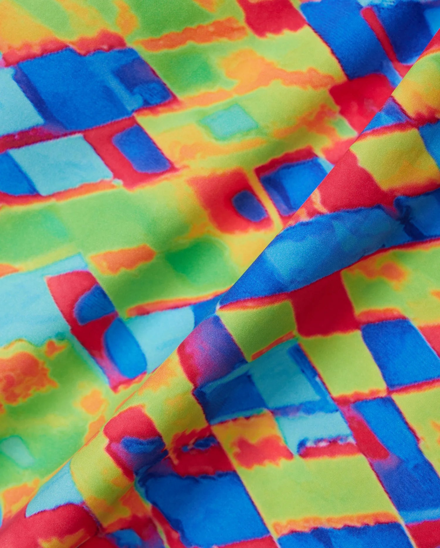 The Psycho Bunny MENS DENNIS ALL OVER PRINT SWIM TRUNK - B6W530C200 showcases a vibrant abstract pattern with geometric shapes in lively hues of red, blue, green, and yellow. The design appears folded, adding an illusion of depth and texture to the quick-dry fabric.