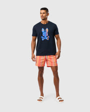 A man stands wearing a black T-shirt with a colorful print on the front and Psycho Bunny MENS ANTON ALL OVER PRINT SWIM TRUNK - B6W542C200. He has short hair and is also wearing white slide sandals. The background is plain white.