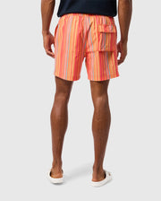 A person stands with their back to the camera, wearing Psycho Bunny MENS ANTON ALL OVER PRINT SWIM TRUNK - B6W542C200 in shades of pink, orange, and blue. The quick-dry trunks have an elastic waistband and a pocket on the right side. The person is also wearing white slip-on footwear.