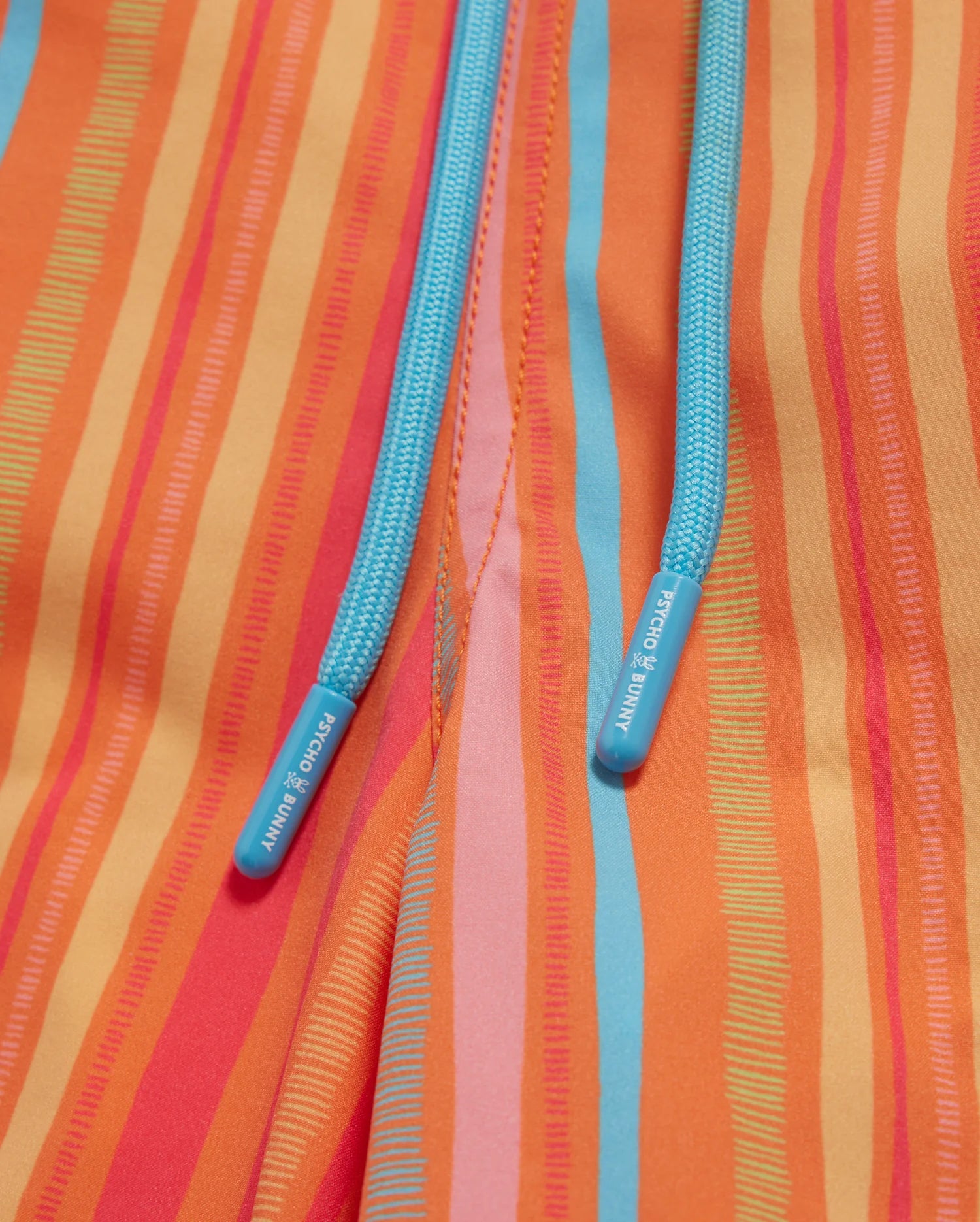 Close-up of a colorful fabric with vertical stripes in shades of orange, yellow, red, and pink. Two turquoise drawstrings featuring the word "Psycho Bunny" at the tips are visible in the center, emerging from seams—perfect for vibrant MENS ANTON ALL OVER PRINT SWIM TRUNK - B6W542C200.