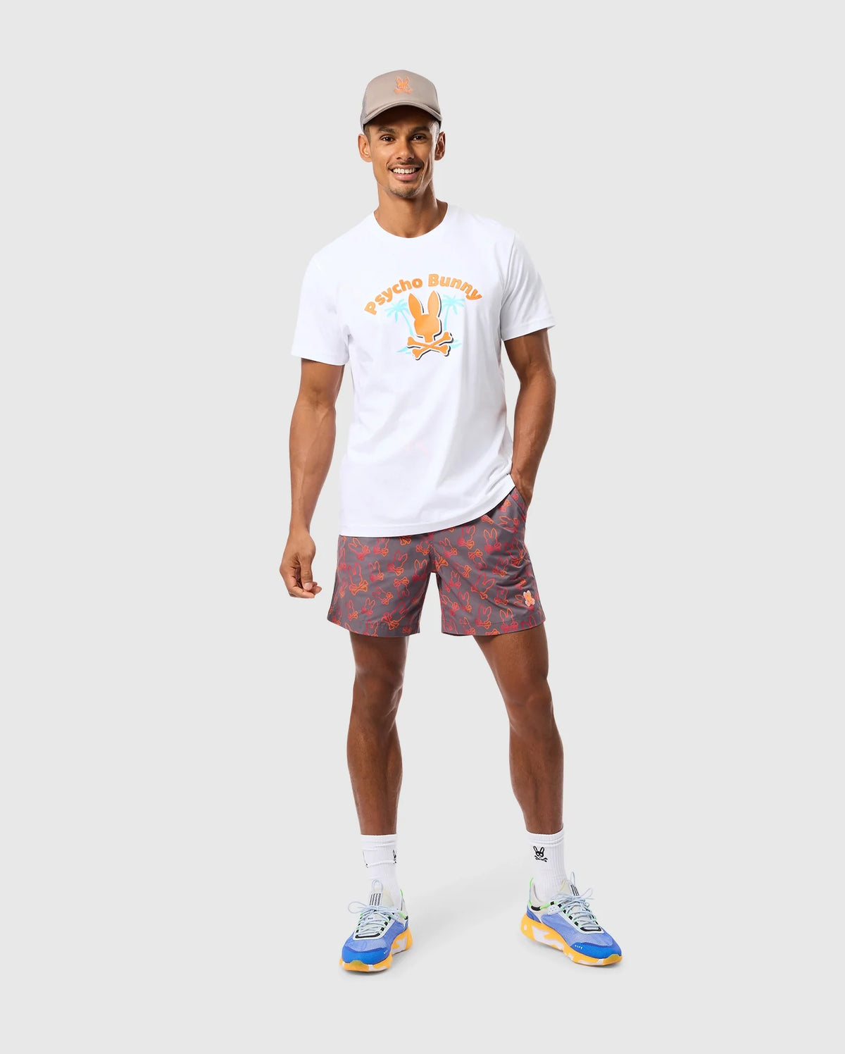 A man is standing against a plain background, smiling. He is wearing a gray cap, a white T-shirt with an orange printed design, and Psycho Bunny's MENS SHELDON ALL OVER PRINT SWIM TRUNK - B6W588C200 featuring a colorful bunny pattern. His outfit is completed with white socks and blue sneakers with yellow soles. He has one hand in his pocket.