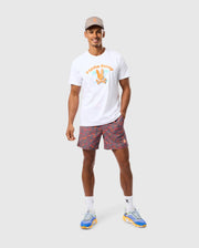 A man is standing against a plain background, smiling. He is wearing a gray cap, a white T-shirt with an orange printed design, and Psycho Bunny's MENS SHELDON ALL OVER PRINT SWIM TRUNK - B6W588C200 featuring a colorful bunny pattern. His outfit is completed with white socks and blue sneakers with yellow soles. He has one hand in his pocket.