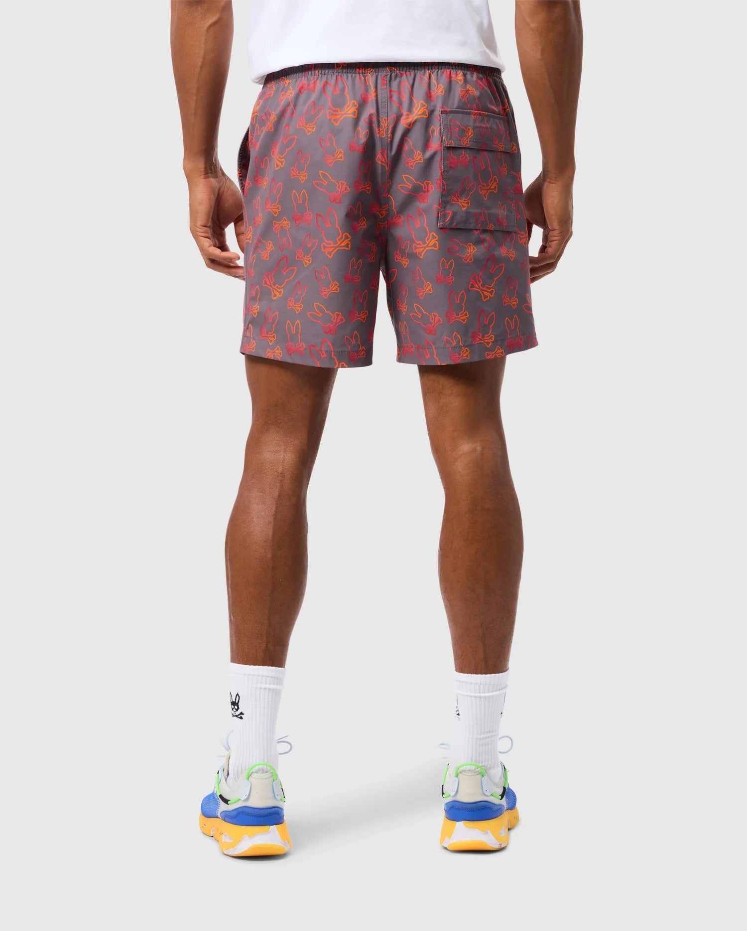 A person is wearing Psycho Bunny's MENS SHELDON ALL OVER PRINT SWIM TRUNK - B6W588C200, characterized by its gray design with an orange bunny pattern and a back pocket on the right side. Complementing the look are white socks with a black logo and colorful running shoes adorned with blue, green, and yellow accents.