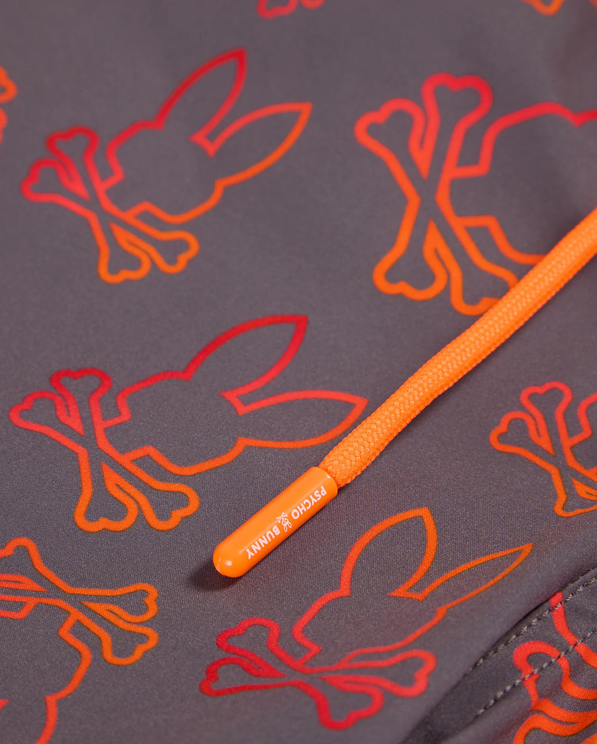 Close-up of the Psycho Bunny Men's Sheldon All Over Print Swim Trunk - B6W588C200, showcasing an intricate bunny pattern with orange and red bunny skull-and-crossbones designs. An orange drawstring with a branded tip labeled "PSYCHO BUNNY" crosses diagonally over the quick-dry grey fabric.