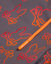 Close-up of the Psycho Bunny Men's Sheldon All Over Print Swim Trunk - B6W588C200, showcasing an intricate bunny pattern with orange and red bunny skull-and-crossbones designs. An orange drawstring with a branded tip labeled "PSYCHO BUNNY" crosses diagonally over the quick-dry grey fabric.