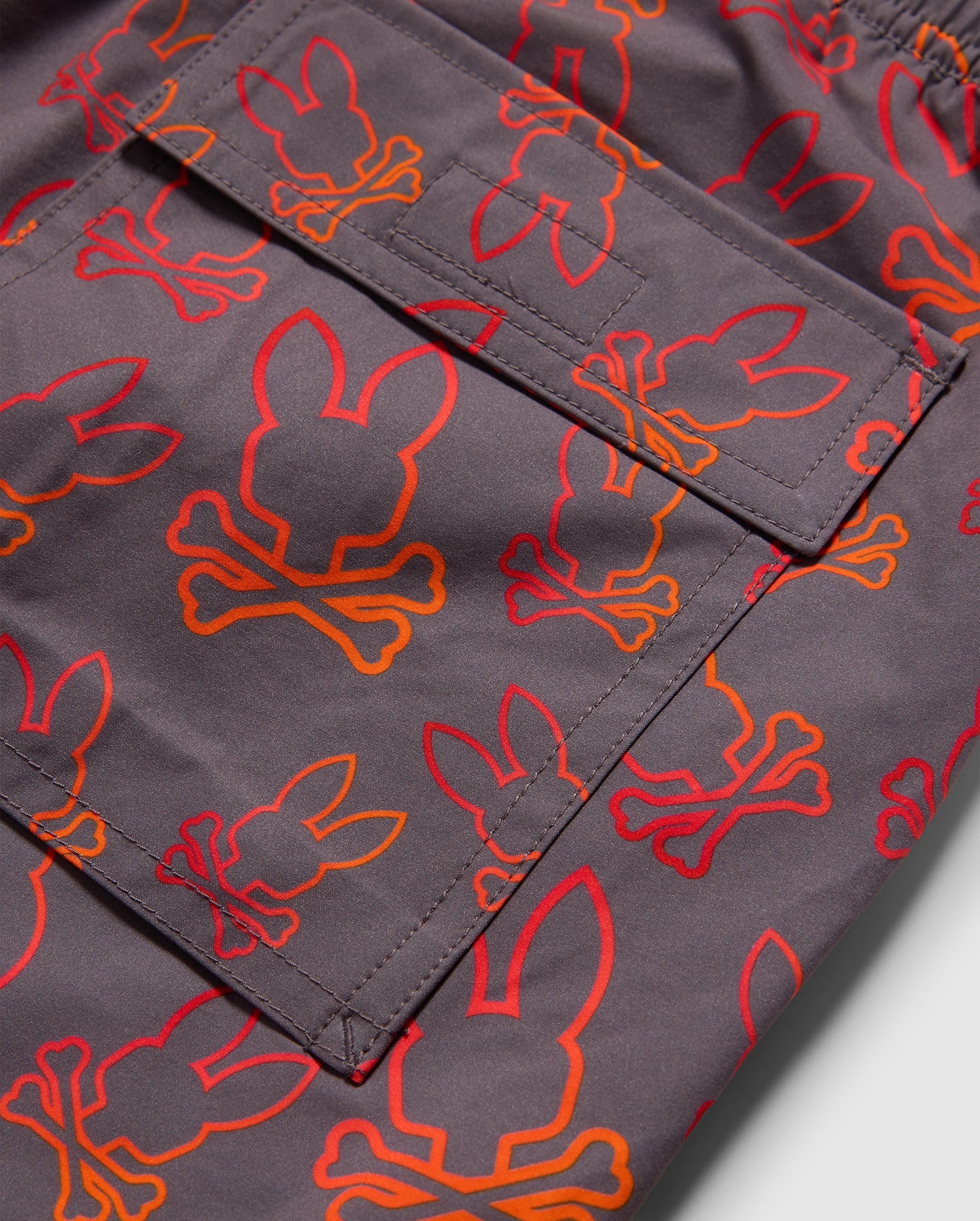 Close-up of the MENS SHELDON ALL OVER PRINT SWIM TRUNK - B6W588C200 by Psycho Bunny showing a gray pocket with an orange and red bunny pattern, featuring bunny heads with crossed bones below them. The design is repeated across the visible portion of the quick-dry fabric.