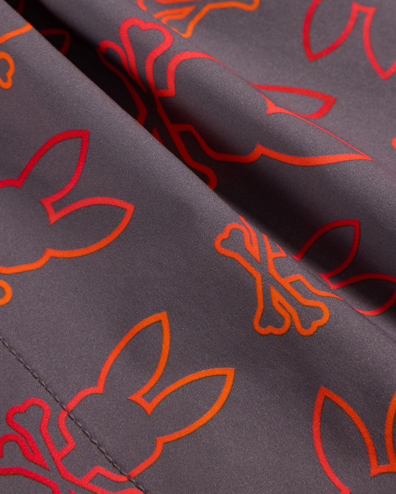 Close-up of Psycho Bunny's MENS SHELDON ALL OVER PRINT SWIM TRUNK - B6W588C200 in grey, featuring a pattern of red and orange outlines of bunny faces and crossed bones. The detailed stitching on the quick-dry fabric is visible, adding texture to the smooth material.