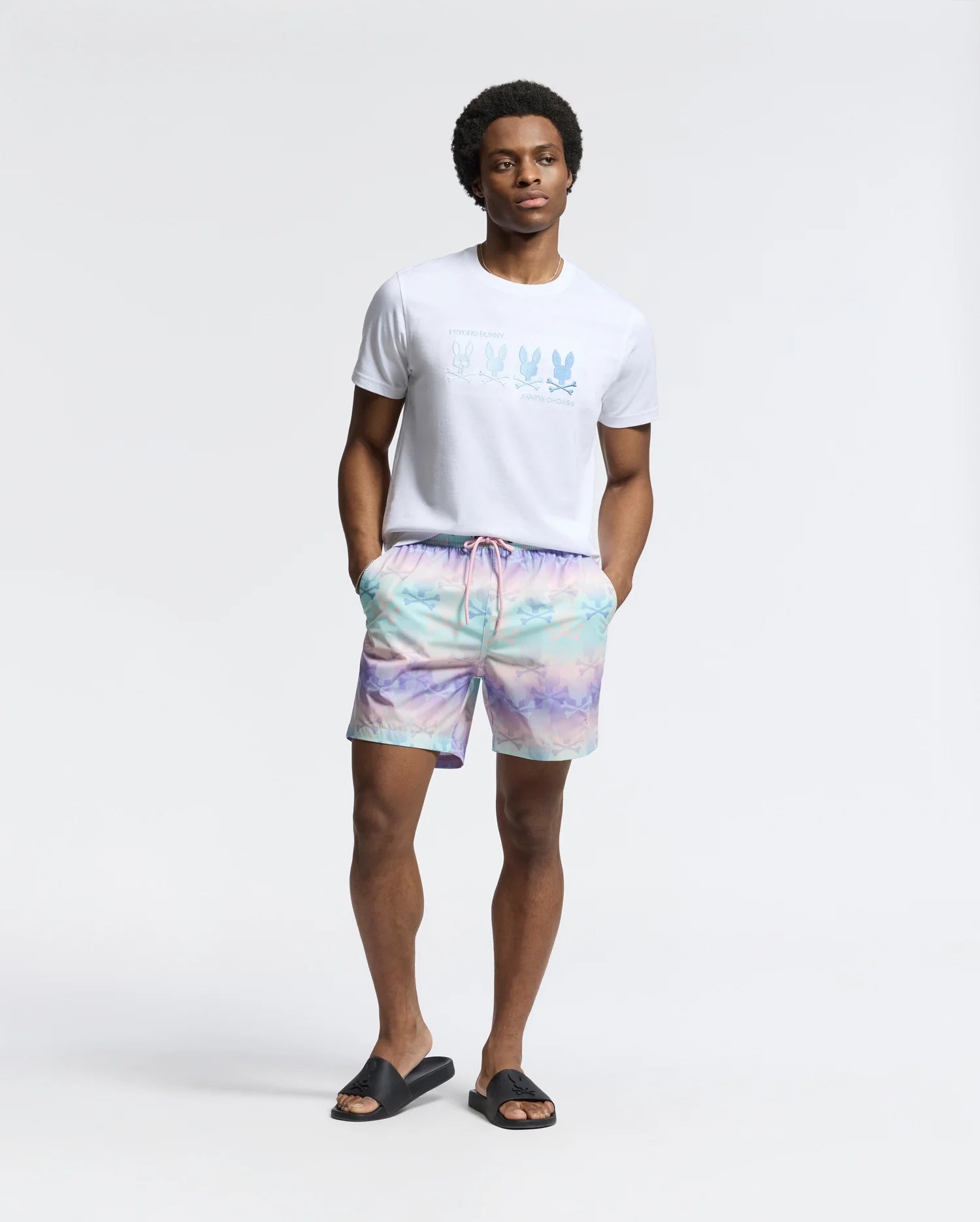 A man exudes beach-bum comfort in a light T-shirt and Psycho Bunny pastel tie-dye MENS FORSTER BUNNY PRINTED SWIM TRUNK. With black slide sandals, he stands casually, hands in pockets, against a plain white background.