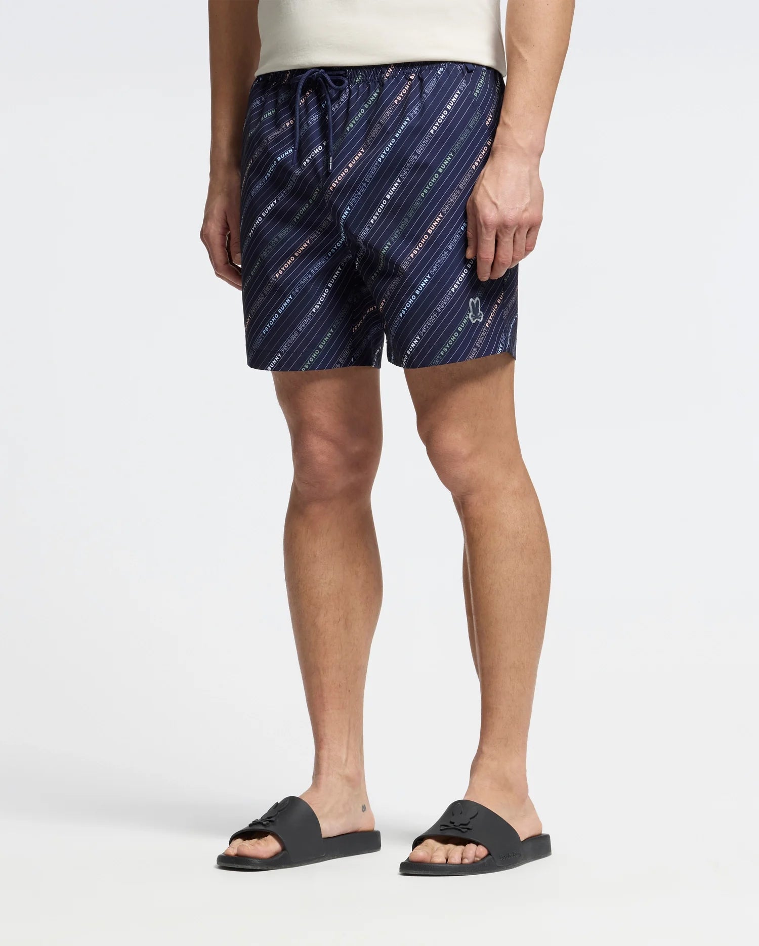 A person in a white t-shirt and Psycho Bunny's MENS RENMARK PB PRINTED SWIM TRUNK (B6W622F200) with diagonal stripes and logo print stands against a light background, paired with black sandals for a casual look.