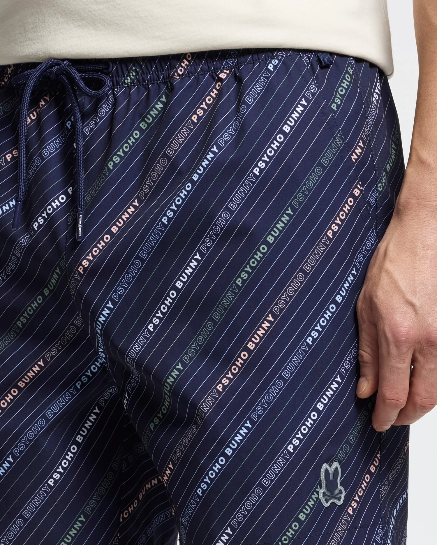 A close-up shows someone in the MENS RENMARK PB PRINTED SWIM TRUNK by Psycho Bunny. The dark blue trunks feature diagonal stripes, a colorful logo print, and a peace sign hand logo on the right leg, all set against a neutral backdrop.