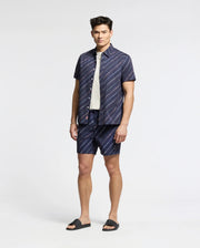 A man stands against a plain background in a relaxed pose, wearing Psycho Bunny’s dark blue and white striped MENS RENMARK PB PRINTED SWIM TRUNK (B6W622F200), paired with a white t-shirt and dark sandals, exuding style as if ready to toss essentials into his waterproof bag.