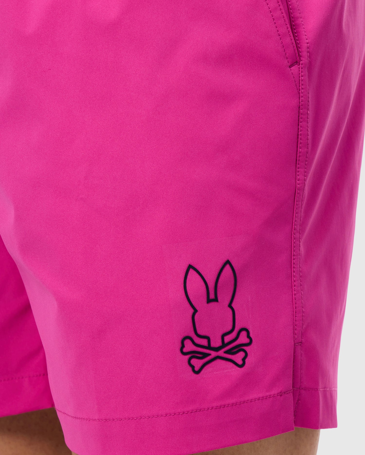 Close-up of a person wearing bright pink, quick-drying MENS PARKER HYDROCHROMIC SWIM TRUNK - B6W646C200 by Psycho Bunny with side pockets. The trunks feature a black outline design logo near the hem showing a rabbit's head above crossed bones, resembling a playful skull and crossbones motif that adds to the unique bunny pattern.