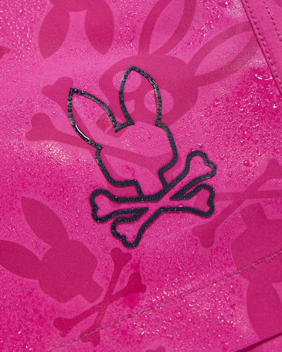 A close-up of a pink fabric with a bunny pattern featuring head silhouettes and crossbones. The water-repellent material has droplets on its surface, suggesting quick-drying Psycho Bunny MENS PARKER HYDROCHROMIC SWIM TRUNK - B6W646C200. Bunnies appear with crossed bones just below their heads, making for a unique design.
