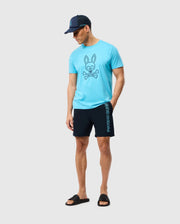 A man wearing a blue t-shirt featuring a bunny and crossbones design from Psycho Bunny, black MENS MELVIN SWIM TRUNK - B6W665C200 with blue lettering on the side, a navy cap, and black slide sandals stands against a plain white background. His hands are by his sides as he looks down.