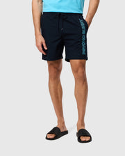 A person wearing black Psycho Bunny MENS MELVIN SWIM TRUNKS - B6W665C200 with "PSYCHO BUN" vertically printed in blue on the left leg, paired with black slide sandals and a light blue shirt. Only the lower half of the person is visible.