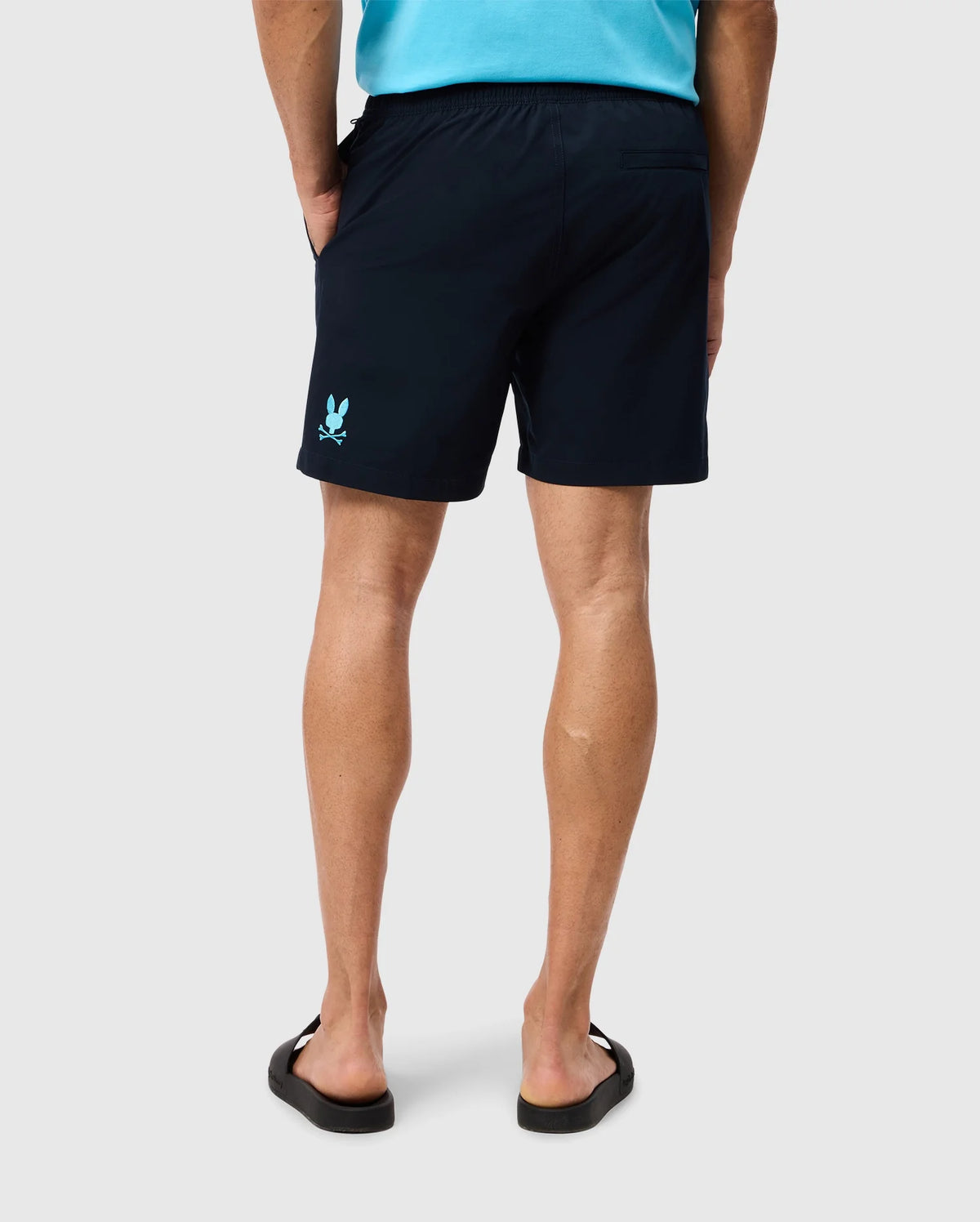 A person wearing a turquoise top and Psycho Bunny's MENS MELVIN SWIM TRUNK - B6W665C200 with a quick-dry outer shell stands with their back facing the camera. The individual has their hands in their pockets and is wearing black flip-flops. The shorts feature a small, light blue logo near the bottom left leg.