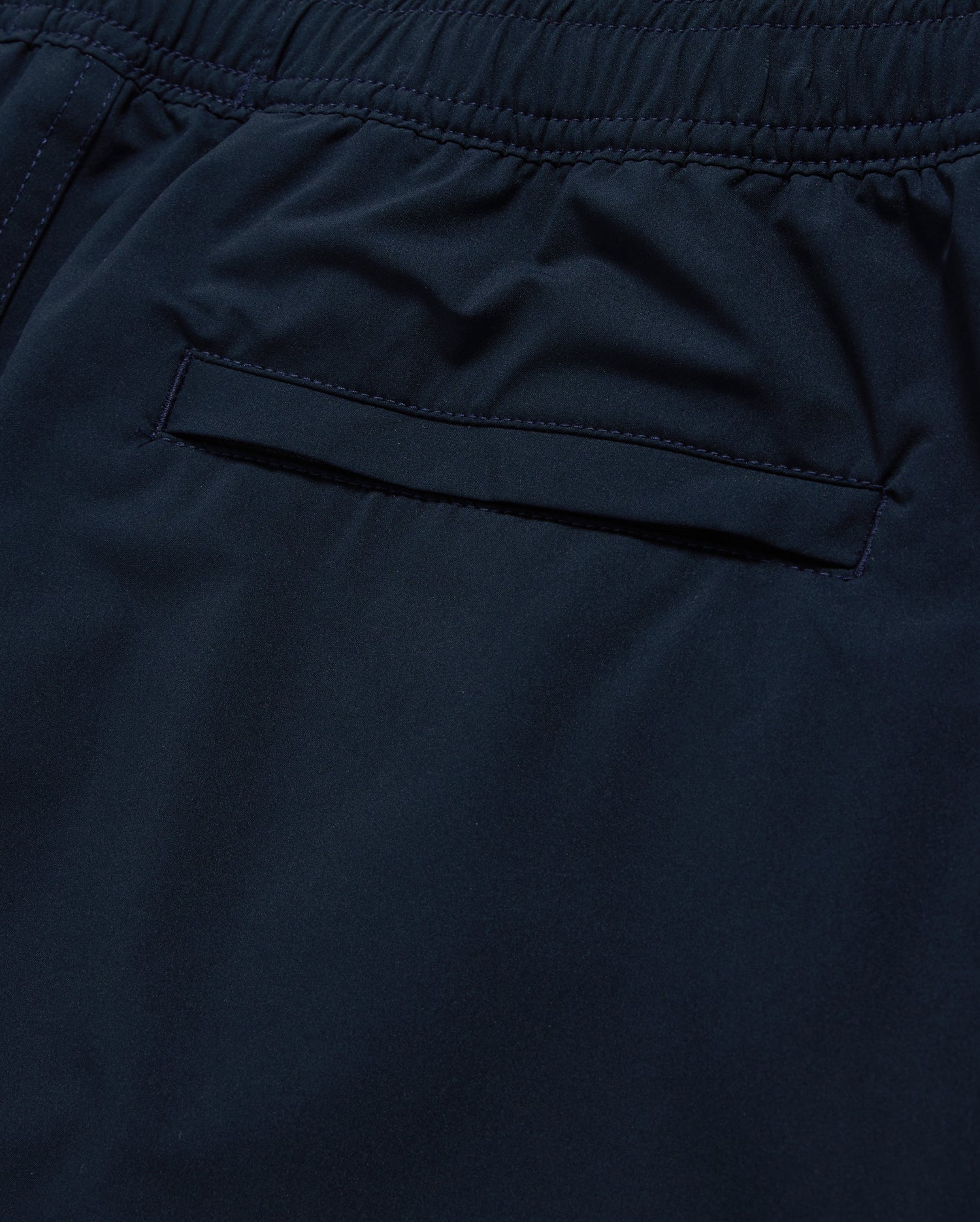 Close-up of a dark blue Psycho Bunny MENS MELVIN SWIM TRUNK - B6W665C200, focusing on a stitched pocket with a concealed zipper. The smooth fabric features a quick-dry outer shell, and the waistband is gathered with elastic and complemented by a drawstring stretch for a comfortable fit. The neat stitching highlights attention to detail in the garment’s design.
