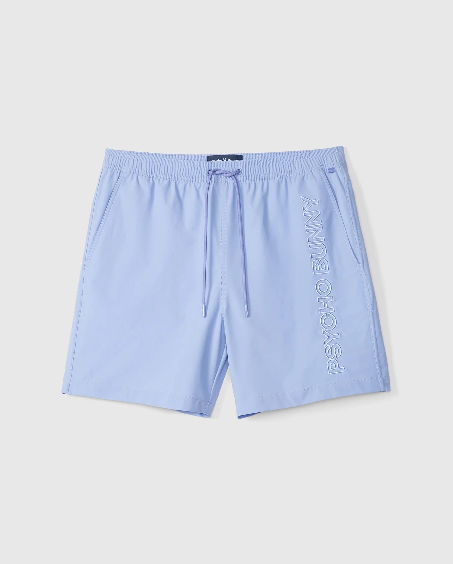 The MENS MELVIN SWIM TRUNK - B6W665C200 by Psycho Bunny features light blue swim trunks with an elastic waistband and drawstring, made from quick-dry textile. Embroidered vertically in white lettering on the right leg are the words "PSYCHO BUNNY." The shorts are laid flat against a white background.