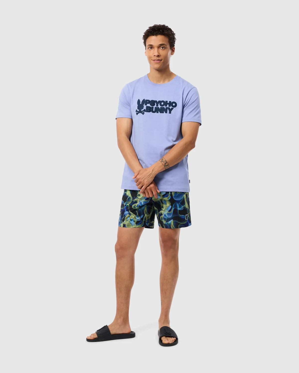 A person stands against a plain background, wearing a light purple t-shirt with a graphic design and Psycho Bunny's MENS GOLDCREEK ALL OVER PRINT SWIM TRUNK - B6W672C200, which features a colorful, wavy pattern. They are also wearing black slide sandals. They are looking at the camera with their hands clasped in front.