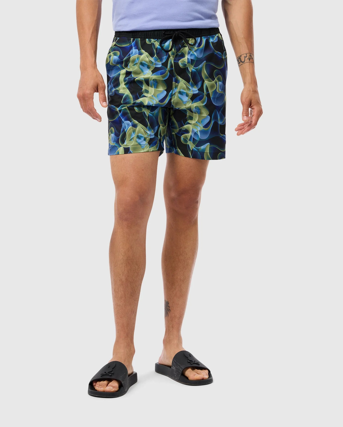 A person is wearing a light purple shirt, vibrant multicolored Psycho Bunny swim trunks with an abstract pattern, and black slide sandals. The shorts, labeled as the MENS GOLDCREEK ALL OVER PRINT SWIM TRUNK - B6W672C200, have a black waistband and pockets. The person's arms are at their sides, revealing a tattoo on their right forearm and another above their left ankle.