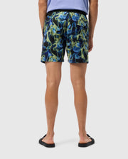 A person is standing and facing away, wearing a light purple T-shirt, Psycho Bunny's MENS GOLDCREEK ALL OVER PRINT SWIM TRUNK - B6W672C200 in blue, green, and yellow abstract patterns, and black flip-flops. The background is plain and light-colored.