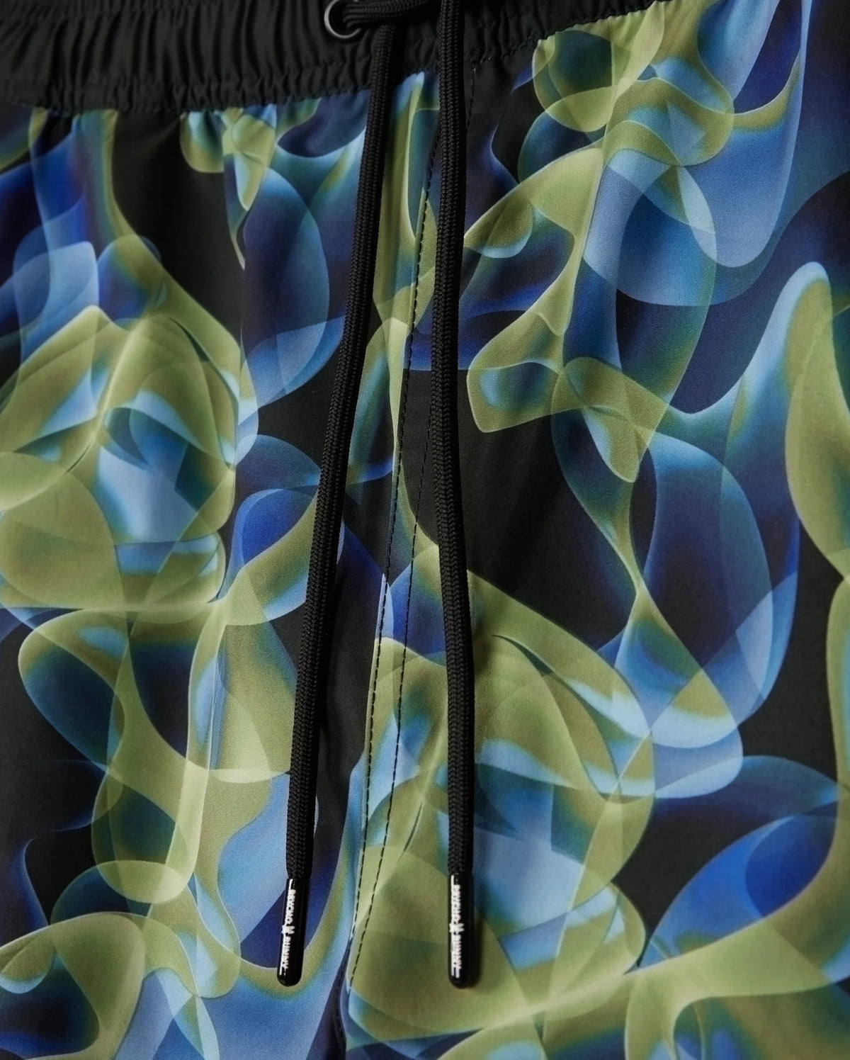 Detailed look at the MENS GOLDCREEK ALL OVER PRINT SWIM TRUNK - B6W672C200 by Psycho Bunny, showcasing a vibrant abstract pattern with swirling shapes in shades of green, blue, and black. The swim trunks feature black drawstrings with small metallic end caps for added style.