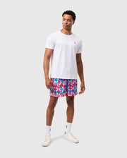 A man is standing against a plain background, wearing a white t-shirt and the Psycho Bunny MENS WALTER LIGHTWEIGHT PRINT SWIM TRUNK - B6W863D200, featuring a vibrant red and blue design. He completes the look with white socks and sneakers. He has short curly hair and is looking slightly to the side, his stance relaxed with one hand by his side.