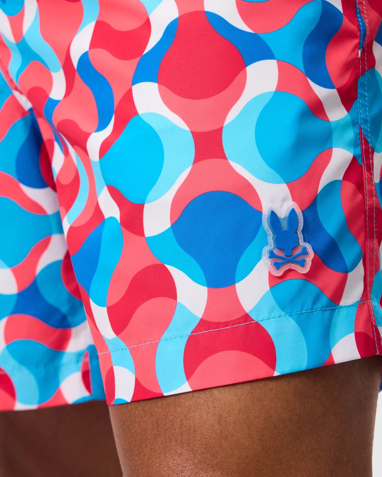 Close-up of a person wearing Psycho Bunny's MENS WALTER LIGHTWEIGHT PRINT SWIM TRUNK - B6W863D200, showcasing the vibrant and bold design. The colorful swim trunks feature a dynamic wavy pattern in blue, red, white, and pink with circular shapes seamlessly blending into one another. Made from 100% polyester, they include a small bunny emblem near the hem against a neutral background.