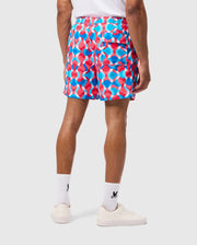 A person is standing with their back to the camera, wearing a white t-shirt and vibrant MENS WALTER LIGHTWEIGHT PRINT SWIM TRUNK - B6W863D200 by Psycho Bunny in blue and red. They complete the look with white socks and sneakers. The background is plain white.