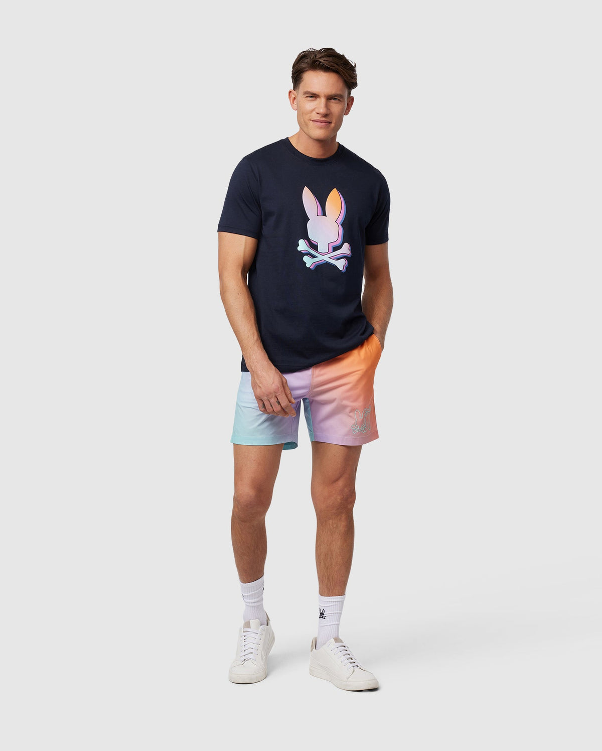 A man wearing a black T-shirt with an HD-printed Bunny logo and Psycho Bunny MENS WINTON PRINTED SWIM TRUNK - B6W915A2PO stands against a plain white background. He is smiling, with one hand in his pocket, and is also wearing white socks and white sneakers.