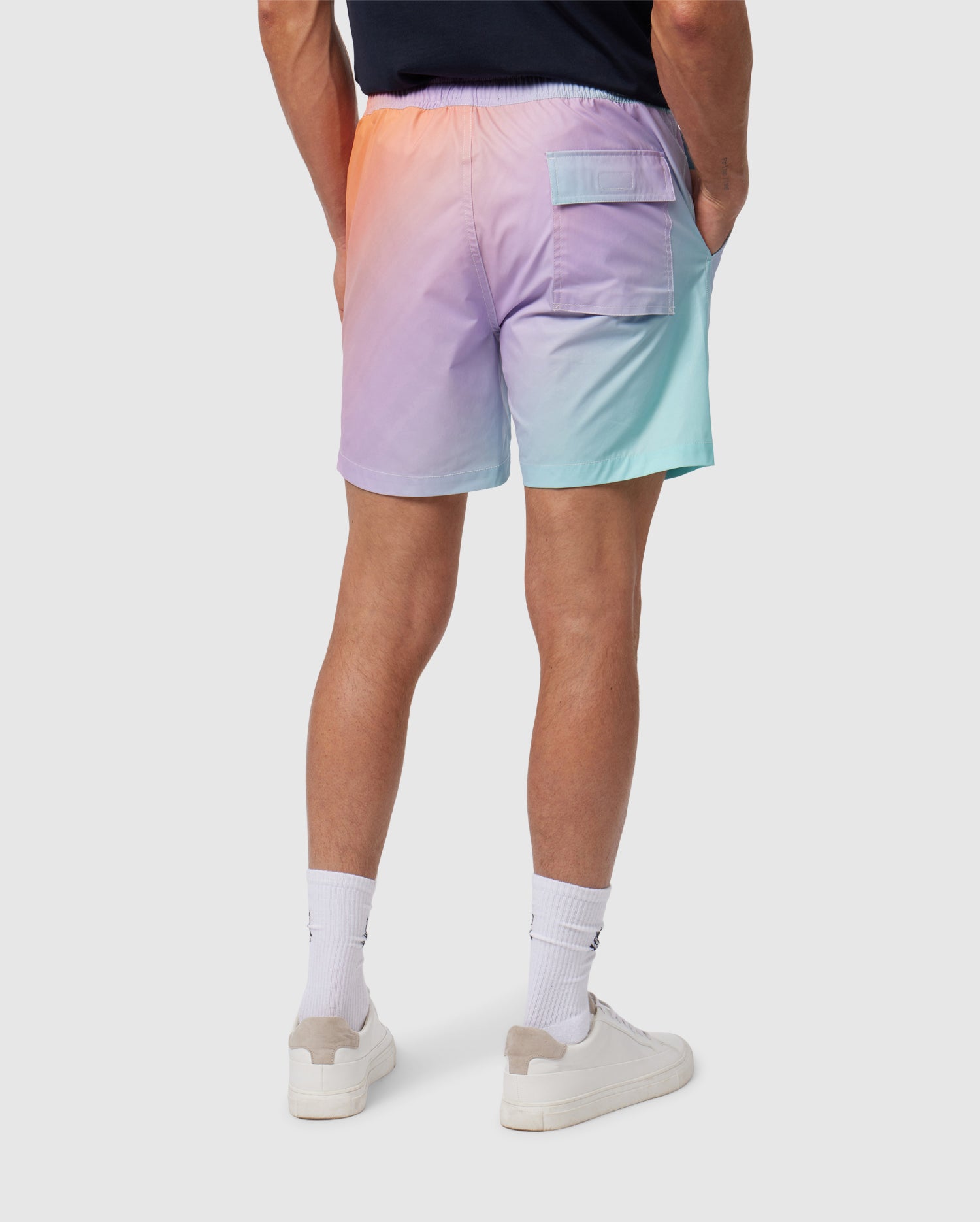 A person is shown from the waist down, wearing Psycho Bunny MENS WINTON PRINTED SWIM TRUNK - B6W915A2PO, which are quick-drying pastel gradient men's swim trunks that transition from orange to purple to light blue. They are also wearing white socks and white sneakers, with hands in the pockets of the shorts. The background is plain.