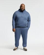 A man faces forward in a blue Psycho Bunny Mens Big and Tall Levy Essential Fleece Hoodie (B9H859D200), paired with matching sweatpants and brown sneakers, set against a plain light background.