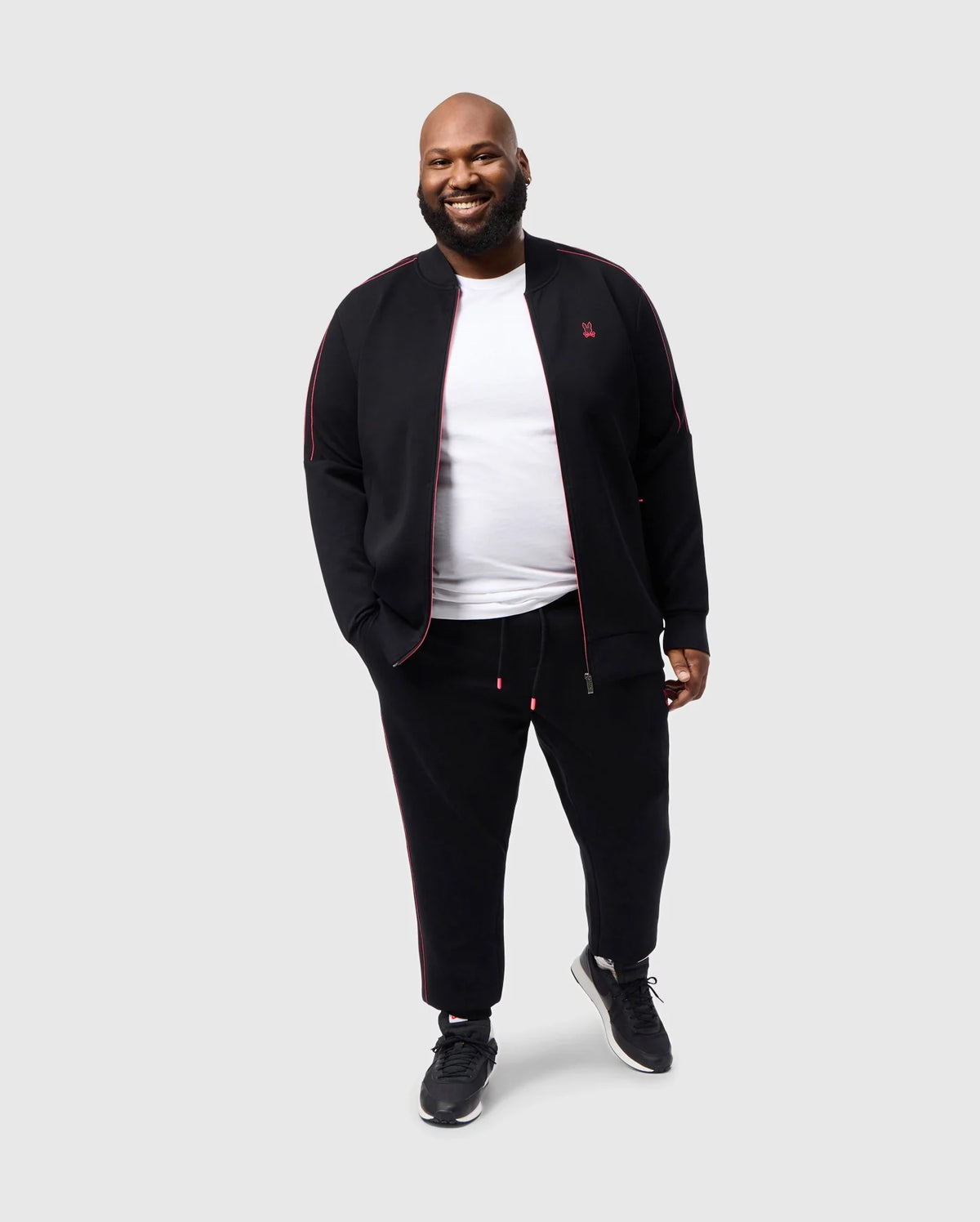 A bearded man smiles against a neutral background, wearing a Psycho Bunny black tracksuit with red accents and a white t-shirt, featuring the BIG AND TALL TRAVIS TRACK JACKET - B9J142D200. Black sneakers complete his casual look.