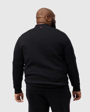 A man stands facing away in a Psycho Bunny BIG AND TALL TRAVIS TRACK JACKET, B9J142D200, against a light gray backdrop. The black cotton-blend jacket features a small red silicone heat-transfer logo near the collar for added style.