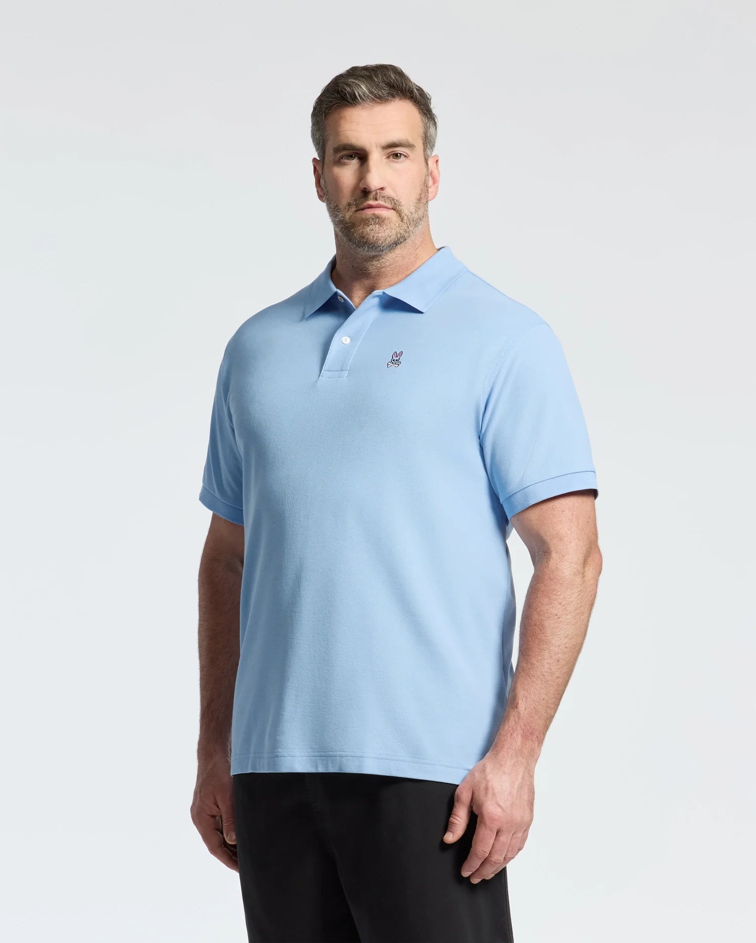 A person in a light blue Psycho Bunny Big and Tall Classic Pique Polo (B9K001CRPC) and dark pants stands against a plain white background.